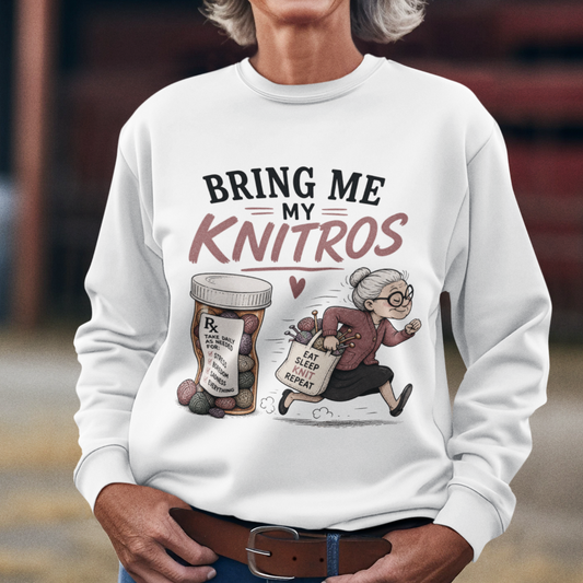 A smiling woman wearing a white long-sleeve graphic sweatshirt, blue jeans, and a white cowboy hat stands outdoors in front of a rustic red barn. The sweatshirt features a cartoon illustration of a knitting machine and yarn spool with the text 'BRING ME MY KNITROS'.